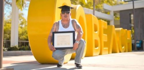 Man in a shirt before a bright yellow Go Beach sign and holding a college diploma