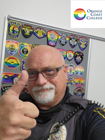 Tim Winer in a public safety uniform in front of a board with patches from various police departments in rainbow LGBTQ+ pride colors